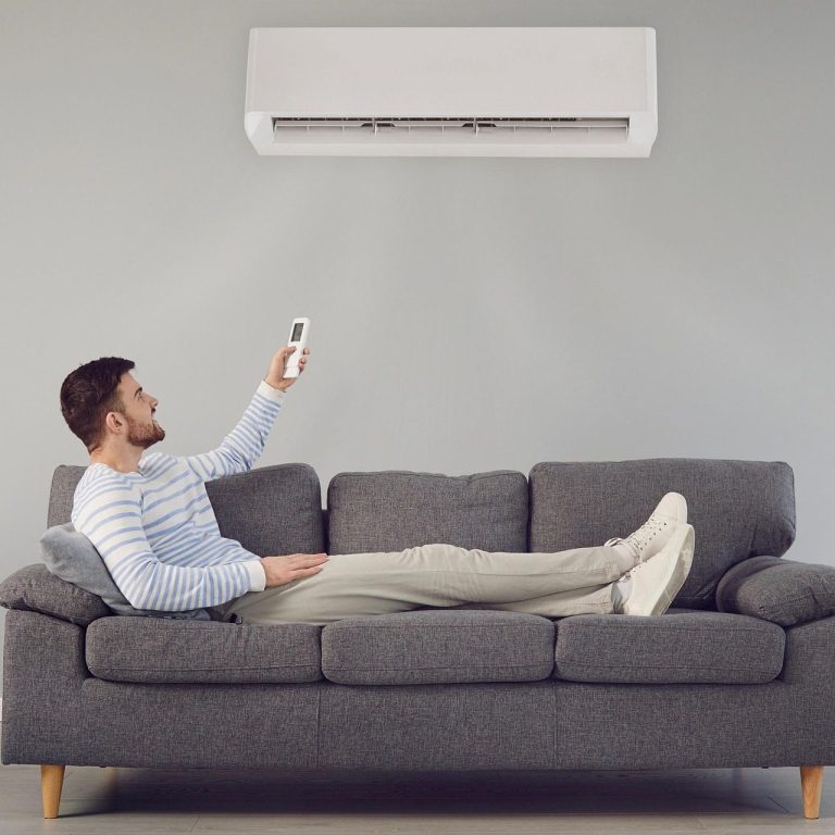 Person entspannt auf einem Sofa und bedient eine Wand-Klimaanlage mit einer Fernbedienung in einem Wohnzimmer.
